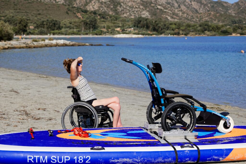 Une femme en fauteuil roulant sur une plage regarde vers la mer, à côté d’un paddle équipé d’un fauteuil tout-terrain bleu, avec des collines verdoyantes en arrière-plan sous un ciel clair.