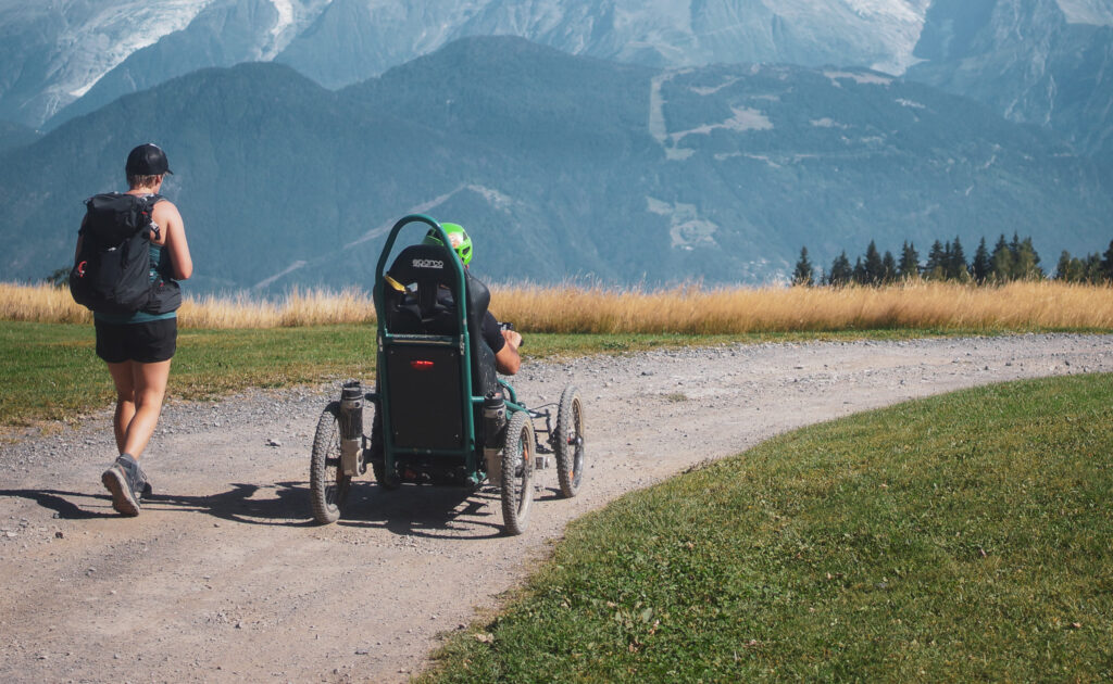 Une femme marche aux côtés d’un homme en fauteuil tout-terrain sur un chemin de montagne, avec les sommets enneigés des Alpes en arrière-plan.