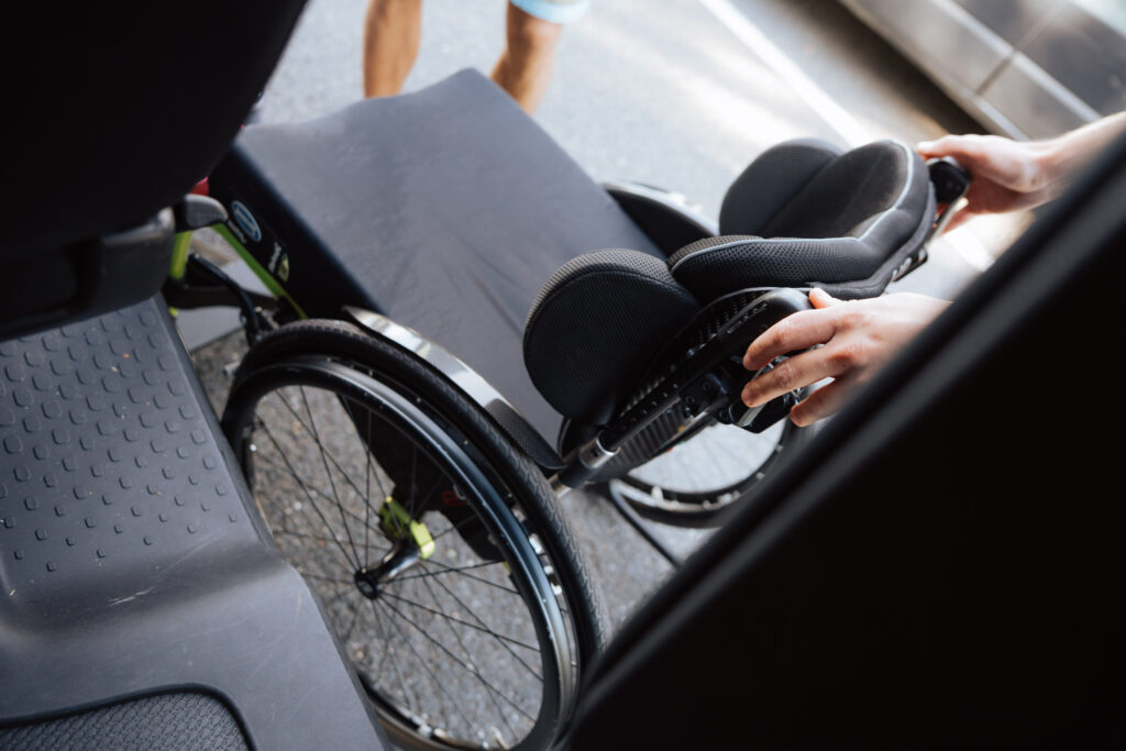 Un fauteuil roulant léger est placé à côté de la porte ouverte d’un véhicule, vue rapprochée sur les mains et les roues.