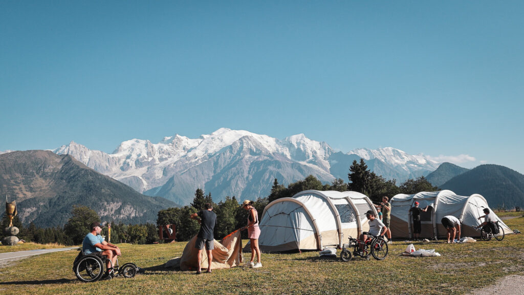 Participants en situation de handicap et valides installent un campement face aux montagnes enneigées des Alpes sous un ciel bleu.
